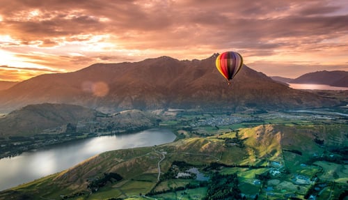 A colourful hot air balloon floats above rolling vineyards and a picturesque countryside in New Zealand.