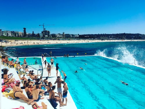 A lively scene with people enjoying a coastal swimming pool, waves crashing over the pool_s edge, and the bustling Bondi beach in the background.