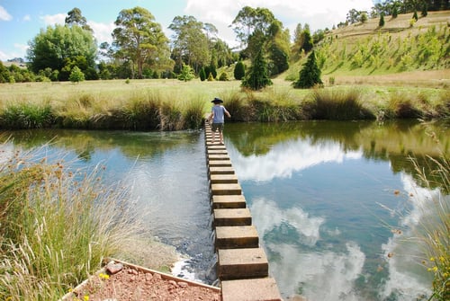 A scene with a child walking on a stepping stone path over a clear, reflective stream, surrounded by greenery and hills.