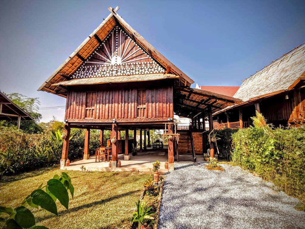 A traditional Lao wooden house on stilts with intricate carvings, surrounded by lush greenery and a garden