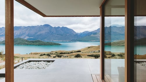 A view from a modern house with large glass windows, overlooking a serene lake and mountains in the distance.