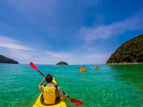 Kayakers paddling through turquoise waters in Abel Tasman National Park, with lush green hills and golden beaches in the background