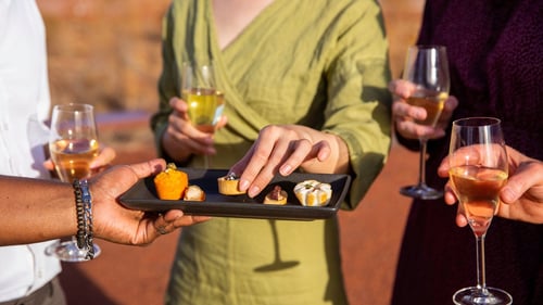 People enjoying wine and appetisers outdoors, with a close-up of hands reaching for food on a tray.