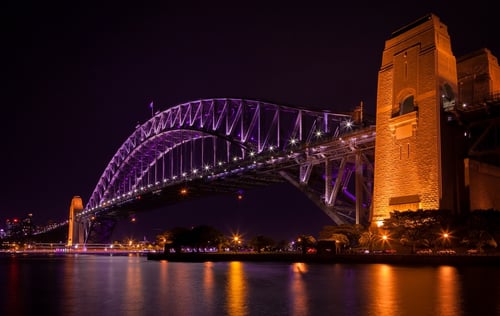 The Sydney Harbour Bridge is illuminated in purple lights against a night sky.