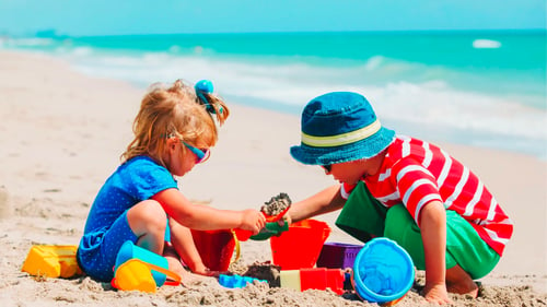 Two children happily build sandcastles and play with beach toys on a sunny beach with turquoise waters.